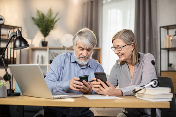 Fototapeta premium Happy elderly spouses holding modern cell phones while resting from distant work on gadgets in home office. Cheerful middle-aged freelancers checking social media via mobiles during short break.