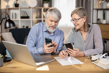 Close up view of mature businesspeople with cell phones looking at each other while staying in comfortable workplace. Senior husband and wife running business together using modern technologies.