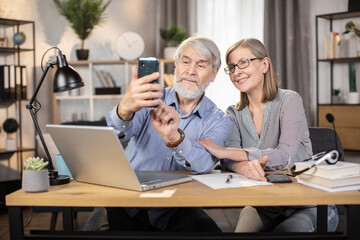 Cheerful mature lady snuggling to attractive senior man while posing for self-picture taken on modern cell phone in home office. Elderly people enjoying comfortable lifestyle while working remotely.