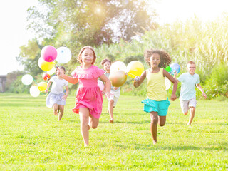 Fototapeta premium Barefoot children running through field with balloons in hands and smiling.