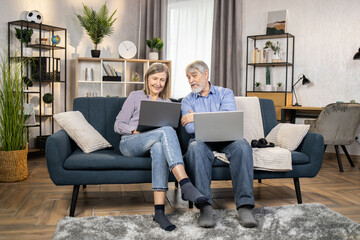Serious mature people in casual clothes using laptops while spending time in cozy environment at home. Elderly family man and wife reading online article on digital screen while sitting on couch.