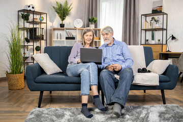 Obraz premium Full length view of relaxed elderly adults watching video on modern laptop while having fun in living room. Happy grandparents relishing kids' birthday party videos shared in social media network.