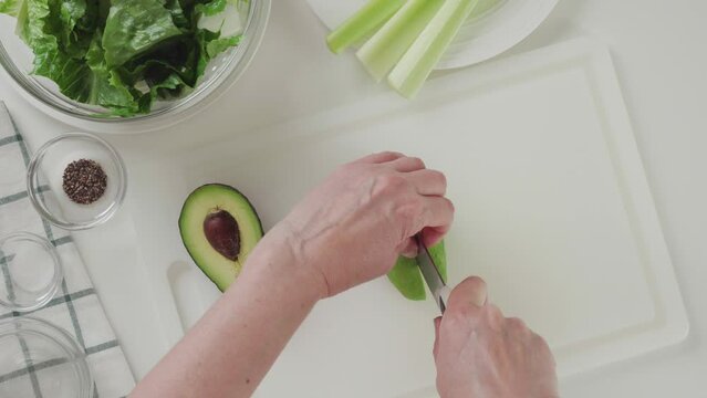 Woman cuts avocado on a white cutting board, view from above. Celery lettuce salad with avocado and lemon dressing recipe
