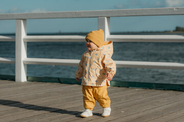 A toddler in a yellow jacket and pants walks on the pier