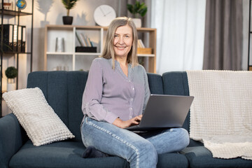 Caucasian woman of 60s resting on soft couch with portable laptop and browsing social media....