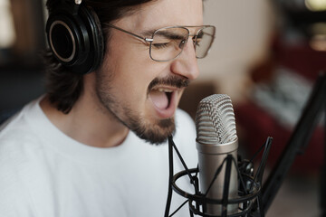 Side view of passionate male singer in headphones sitting at table near microphone and recording...