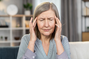 Frustrated senior lady suffering from strong sudden migraine and dizziness while sitting on couch. Sick aged woman in striped shirt massaging temples with fingers indoors. Concept of healthcare.