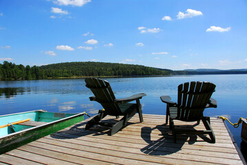 Naklejka na ściany i meble Two adirondack wooden chairs on dock facing a blue lake with clouds reflections