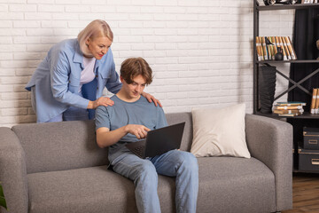 Adult son and middle-aged 40s mother sitting on couch in cozy living room holding on lap notebook...