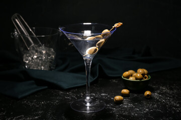 Glass of tasty martini with green olives and ice cubes on dark background