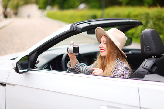 leisure, road trip, travel concept - happy girl driving in cabriolet car and taking picture by film camera