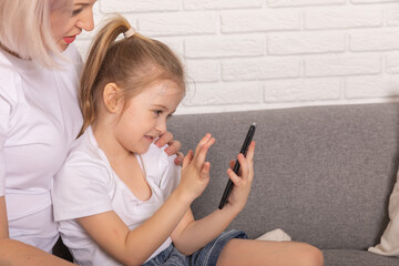Close up happy mother with little daughter kid using smartphone sitting on couch at home, smiling caring mom and adorable girl child looking at device screen, chatting online, watching cartoons