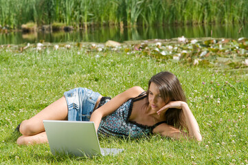 cute brunette in casual dress laying down on the grass and working on the computer