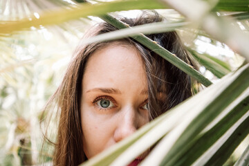 Portrait of young brunette woman among palm leaves. A girl hiding face behind the leaves on a sunny...