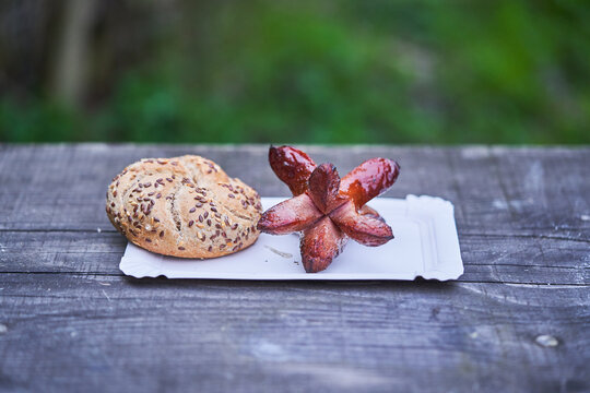 Grilled traditional small pork sausage over open campfire and served with rustic wholegrain gread roll or bun on paper plate. Typical specialty from Czech republic served as a fast food or party snack
