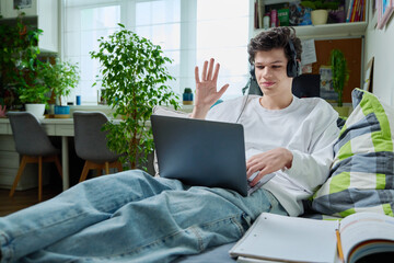 Young guy in headphones using laptop for video communication, lying at home