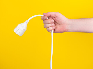 A hand holds a plug to which a white cable is attached. No face, yellow background. 
