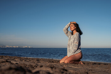 beautiful brunette woman in a sweater and swimsuit sits on the sand by the sea enjoys the environment