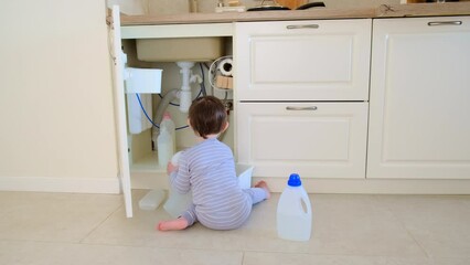 A child is playing with chemical cleaning products under the sink in the kitchen. Baby holds bottles with detergent. Kid aged about two years (one year nine months)