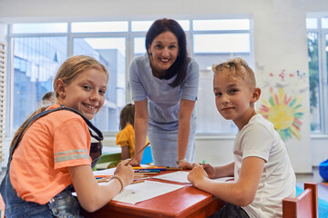 Creative kids during an art class in a daycare center or elementary school classroom drawing with female teacher.