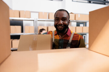 Smiling african american manager working at customers order, putting products in cardboard boxes, preparing products for shipping. Employee wearing industrial overall while carrying packages