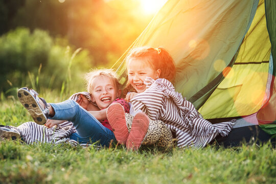 Two little girls sisters sitting on the green grass next to camping tent entrance, cheerfully laughing about jokes they told to each other. Careless childhood and outdoor activities concept photo.