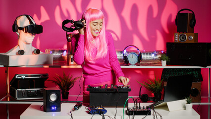 Happy artist with pink hair and blouse mixing sounds at professional mixer console, listening music into headset during party. Asian dj performer dancing and having fun in club at evening.