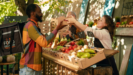 African american deliveryman wearing backpack waiting for order at farmers market counter. Team of farmers delivering fresh organic fruits and vegetables to customers, small business owners.