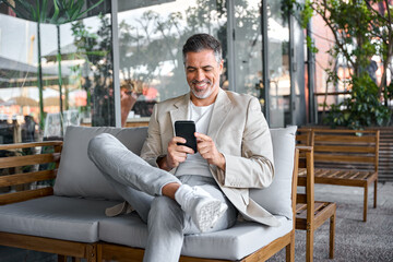 Happy smiling relaxed mid aged business man, mature professional businessman entrepreneur sitting in outdoor cafe holding smartphone using mobile phone digital technology apps. Authentic shot