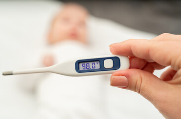 A hand holds a thermometer showing the temperature on the background of a blurred baby lying on the bed
