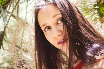 Portrait of a young beautiful enigmatic woman in a playful mood on nature. A girl with cute baby face and blue eyes in tropical garden. Human and nature unity. Straight gaze, look. Good spirits youth.