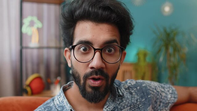 Oh my God, Wow. Young indian man surprised looking at camera with big eyes, shocked by sudden victory, good win news, celebrating. Portrait of excited amazed hindu guy at home apartment room on sofa