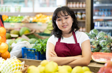 Smiling young saleswoman posing happily in front of counter in grocery store with large assortment