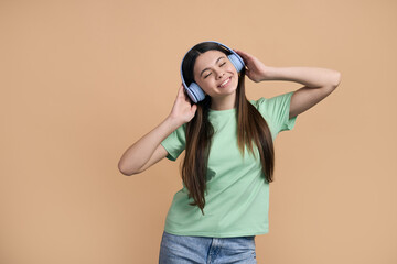 Charming teenage girl putting on blue wireless headphones, listening to music over beige background