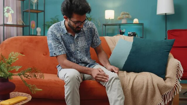 Young indian man suffer from painful severe knee joint ache, leg muscles, could not walk. Portrait of hindu guy massaging his knees illness cramps rheumatism at home room on sofa. Health care problem