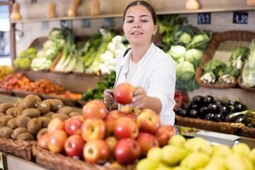 Portrait of positive interested young woman buying fruits in greengrocery store, choosing ripe apples arranged in woven box on produce display..