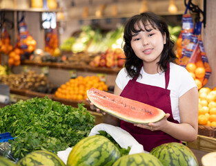 Smiling young saleswoman holding a slice of watermelon at the counter in big grocery store