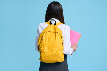 Back view. Smart school girl with yellow backpack standing back to camera, isolated blue background