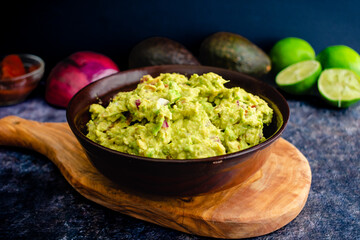 Fresh Guacamole in a Wooden Bowl with Ingredients in the Background: Homemade guacamole with avocados, limes, onion, cilantro, and chipotle peppers