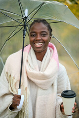 African woman with coffee and umbrella looking at camera and smiling in autumn.