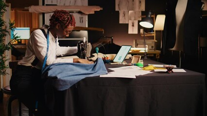 Designer working on sewing machine to manufacture clothing items at workstation, professional tailor. Young couturier using tailoring equipment to create custom made clothes in atelier.