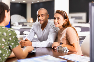 Young happy cheerful positive hispanic couple consulting with furniture saleswoman when choosing new mattress in store