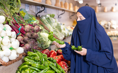 Attentive young Muslim woman purchaser choosing peppers at the counter in large grocery store