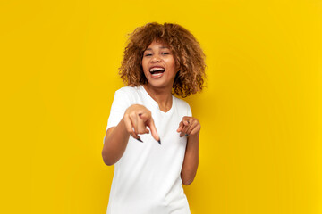 hey you. young curly american girl with braces pointing at you and laughing on yellow isolated background - obrazy, fototapety, plakaty
