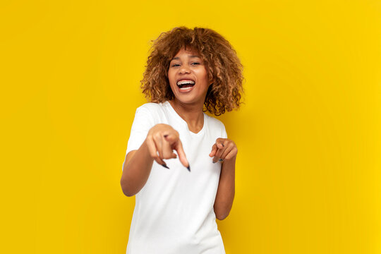 hey you. young curly american girl with braces pointing at you and laughing on yellow isolated background