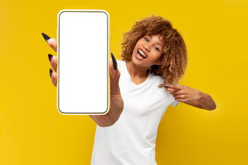Fototapeta na wymiar young curly american girl with braces shows blank smartphone screen on yellow isolated background