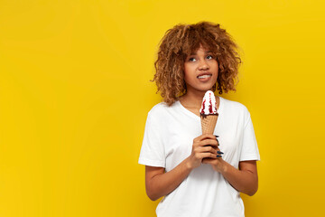 young curly american girl with braces eats sweet ice cream with jam and dreams on yellow isolated background