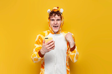 young curly guy in funny giraffe pajamas uses smartphone and rejoices in victory on yellow...