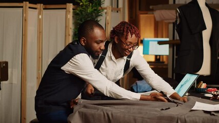 Tailoring partners cutting fabric to design clothes, using scissors to prepare garment for custom made tailored suit. Tailor and assistant working in atelier workshop. Tripod shot.