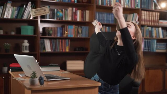 A young woman is sitting at her workplace, she is in a good mood, happy and joyful, when suddenly money pours down from above and she happily collects it.